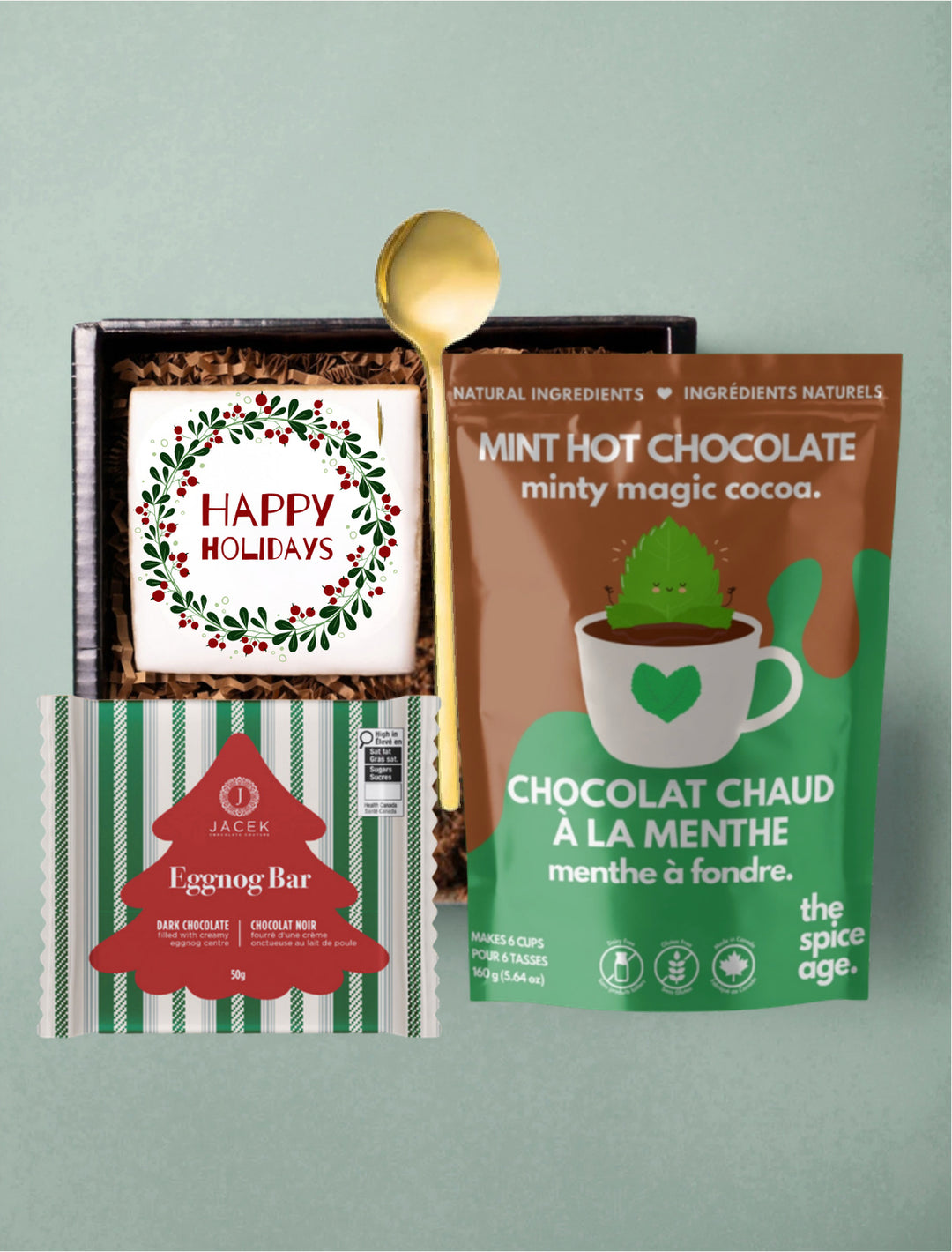 Happy Holidays gift selection with a sugar cookie, JACEK Eggnog Bar, The Spice Age mint hot chocolate and a gold spoon.